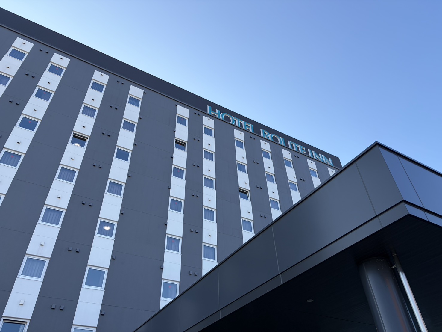 ホテルルートイン砺波インターの外観。灰色の壁に白い窓の列が並び、青空に社名の看板が見える